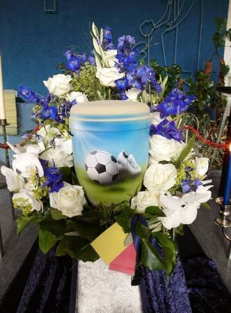 Urne mit Fußballmotiv, umgeben von weißen Rosen und lila Blumen, auf Trauerfeier.