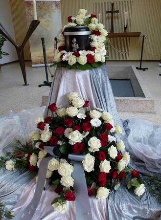 Urne auf Trauerfeier mit Blumengestecken aus weißen und roten Rosen vor Altar.