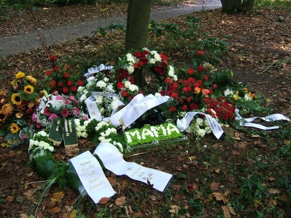 Blumen und Trauerkränze mit Trauerschleifen liegen auf einem Grab im Wald.