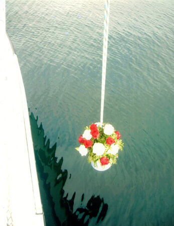 Blumengesteck mit roten und weißen Rosen wird an einem Seil über ruhigem Wasser abgesenkt.