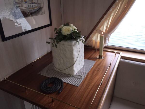 Blumengesteck, Kerze und Seil auf Holztisch in Yachtkabine mit Fenster und Gardine.